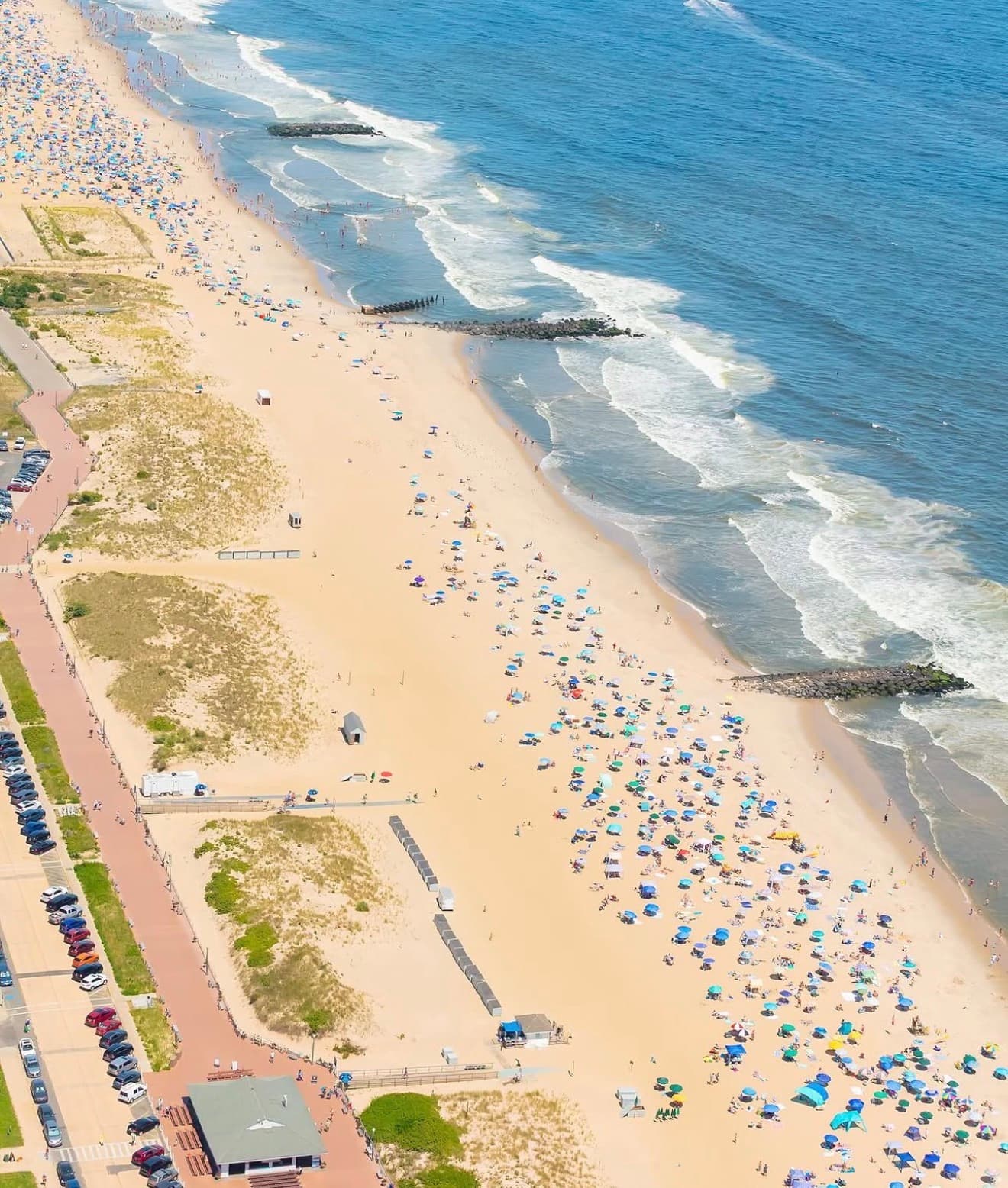 Aerial view of Ocean Grove Beach in New Jersey, showing the wooden boardwalk along the left side, dunes with split-rail fencing, cars parked in rows along Ocean Avenue, rock groins extending into the Atlantic, and crowds of beachgoers with colorful umbrellas on a summer day