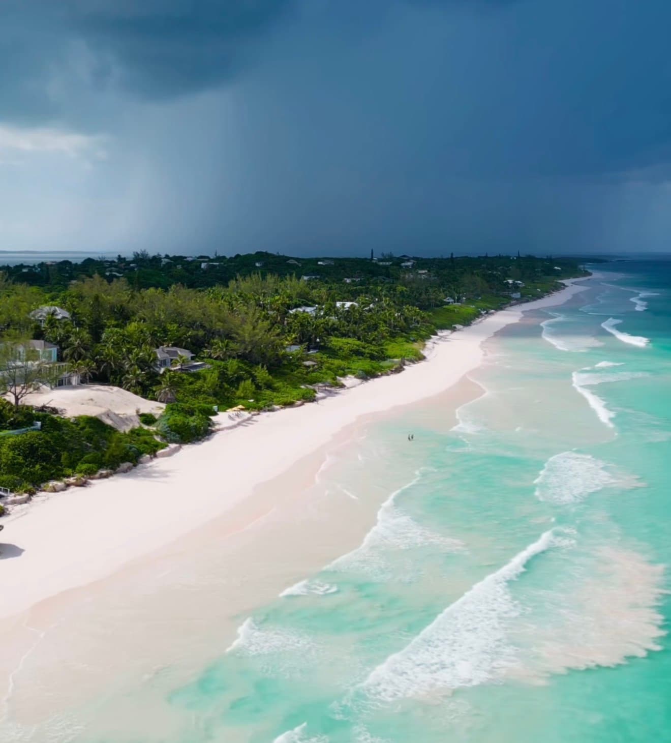Aerial view of the Pink Sand Beach on Harbour Island in the Bahamas, showing a long curve of pale pink sand, turquoise Atlantic water, low-lying tropical vegetation and houses behind the beach, under a dramatic cloud-heavy sky