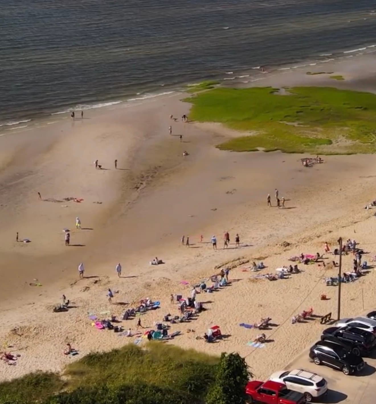 Aerial view of Skaket Beach in Orleans on Cape Cod Bay, showing sunbathers on the sand, calm turquoise water, patches of salt marsh, and cars in the parking lot