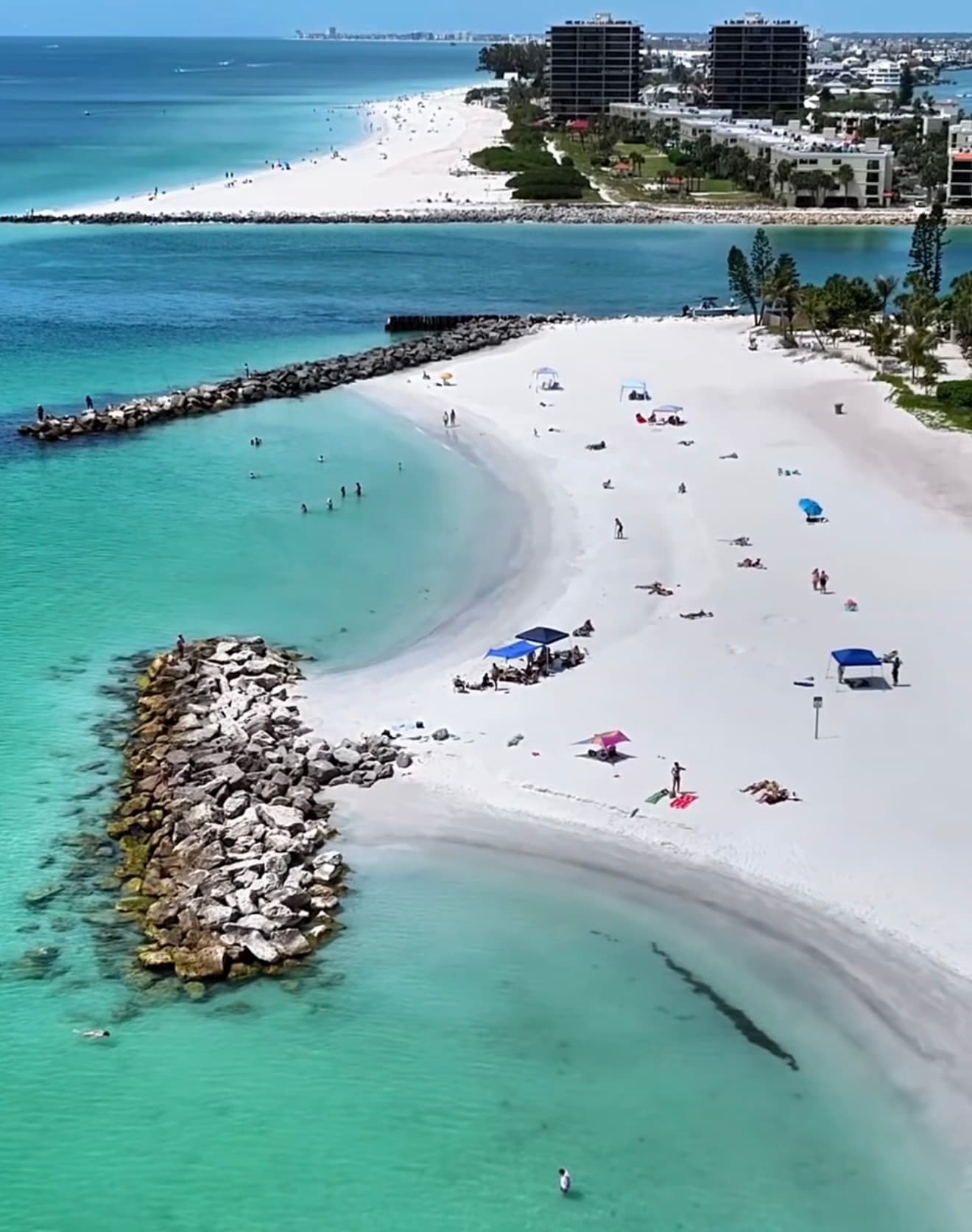 Wide view of Upham Beach in St Pete Beach, Florida, showing freshly renourished pale sand, one of the black rock T-groins extending into the Gulf of Mexico, Blind Pass and the southern end of Treasure Island visible across the water, with beachfront condo towers behind the beach