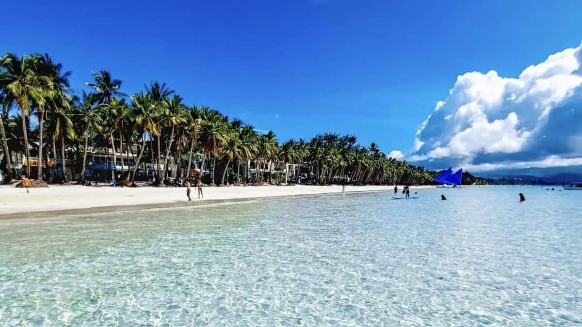 White Beach, Boracay: crystal-clear shallow turquoise water lapping the powder-white sand, with the palm-lined Station 2 beachfront of resorts and a blue paraw sail off to one side