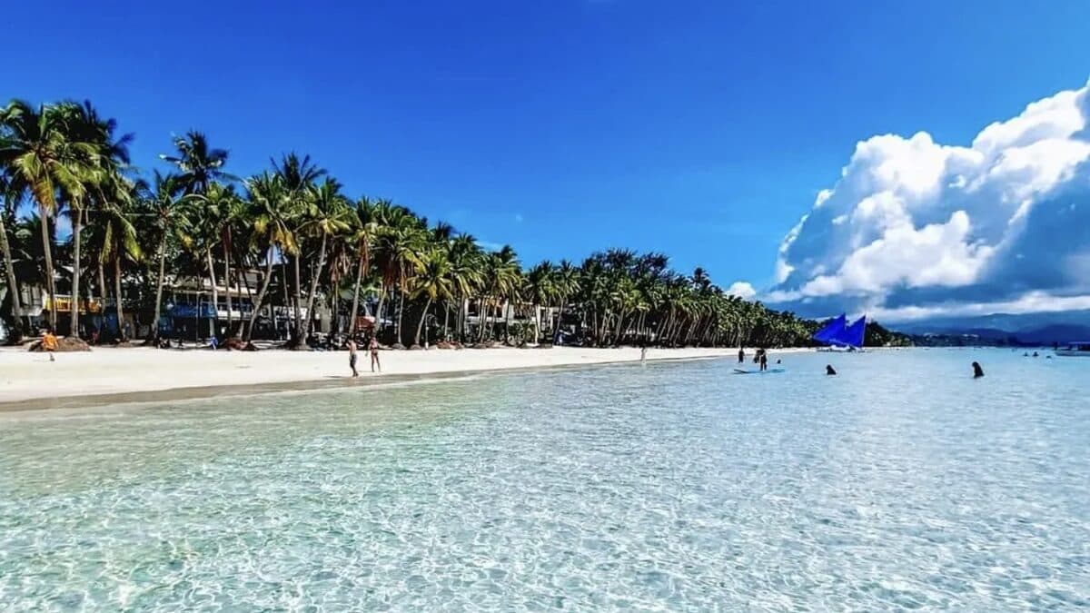 White Beach, Boracay: crystal-clear shallow turquoise water lapping the powder-white sand, with the palm-lined Station 2 beachfront of resorts and a blue paraw sail off to one side