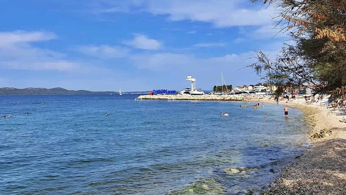 Borik beach in Zadar, the pebble shoreline curving toward the D-Marin Borik marina and its diving tower, with swimmers in the clear shallow water and pines along the shore