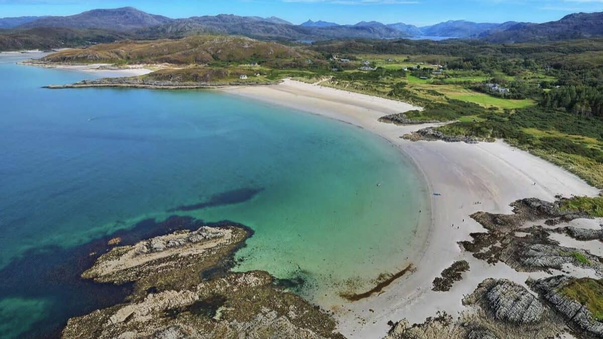 Aerial view of Camusdarach Beach near Morar, the curving white shell-sand crescent and green-turquoise water with rocky outcrops, machair and the Highland hills behind