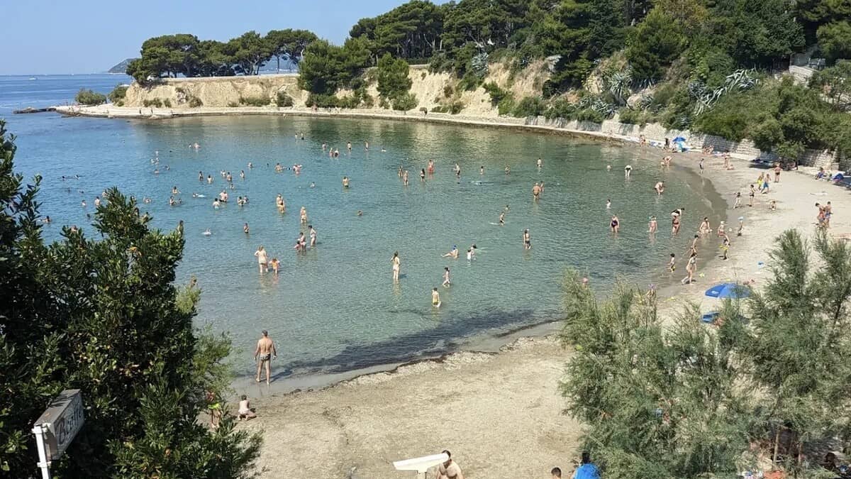 Firule beach in Split, the shallow sandy bay busy with waders and swimmers in the calm clear water, backed by a low pine-topped cliff