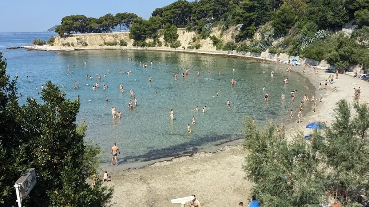 Firule beach in Split, the shallow sandy bay busy with waders and swimmers in the calm clear water, backed by a low pine-topped cliff