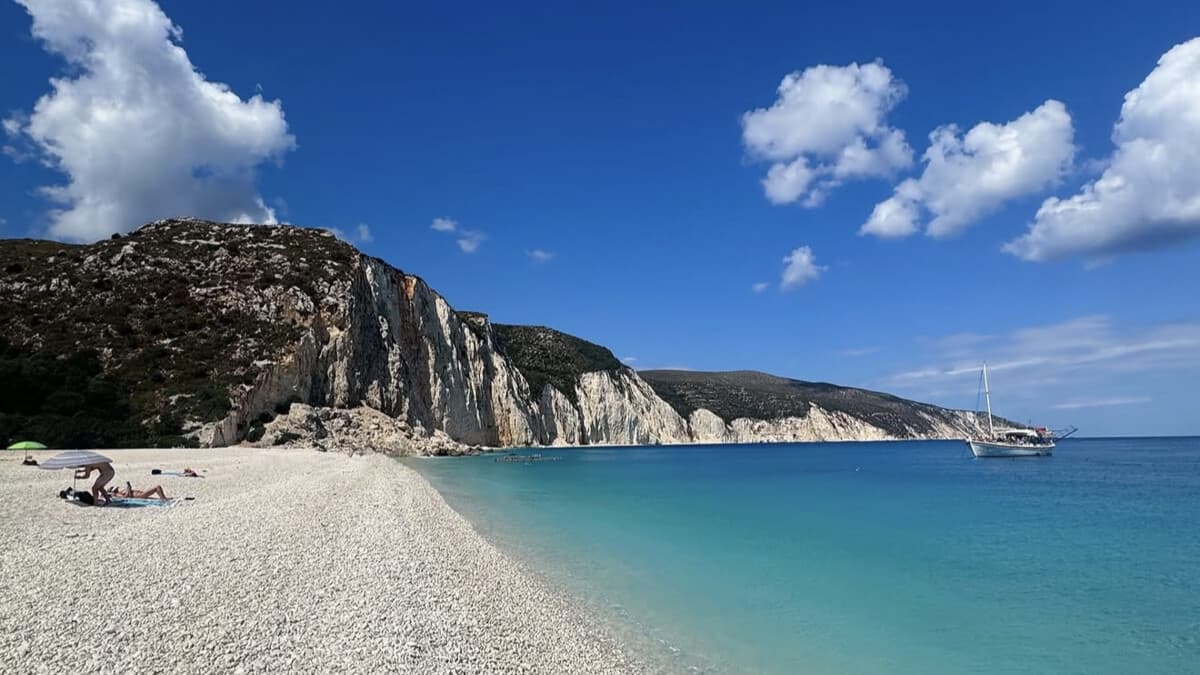 Fteri Beach on Kefalonia, a white-pebble beach beneath dramatic pale cliffs with crystal-clear turquoise water and a boat anchored offshore