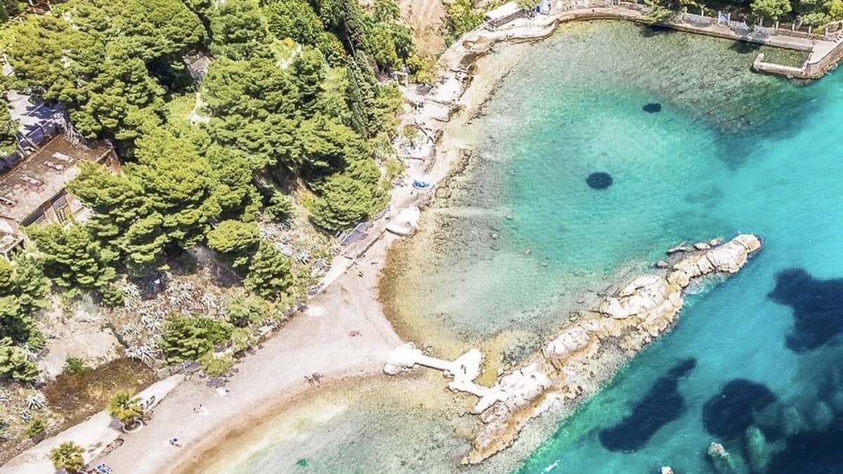Aerial view of Jezinac beach below Marjan in Split, a small pebble cove of clear turquoise water with a rocky breakwater, backed by the pine-covered slope