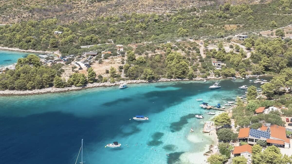 Aerial view of Labadusa beach on Ciovo island near Trogir, a pebble bay of clear turquoise water with a beachfront restaurant, anchored boats and pine-covered slopes