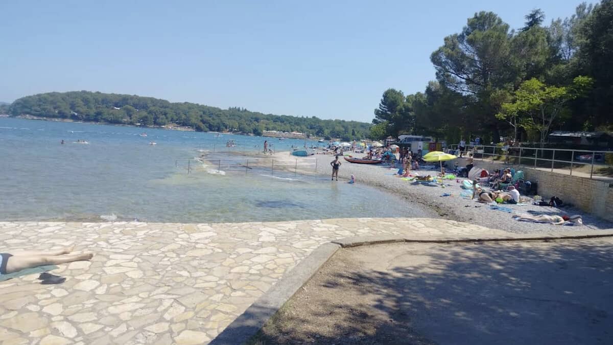Lanterna beach near Porec, the pebble resort shore and paved promenade busy with sunbathers and umbrellas, backed by pines on the Lanterna peninsula