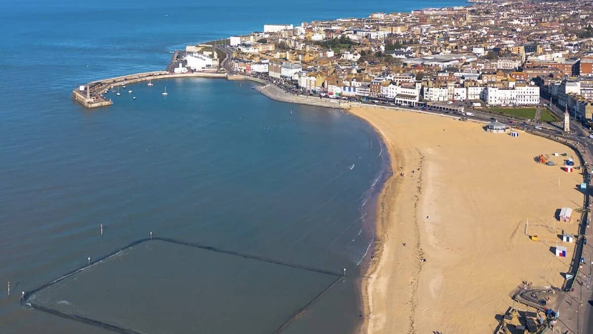 Aerial of Margate Main Sands in Kent, a broad curve of golden sand beside the seafront town, with the old harbour arm curving out to the left and a rectangular tidal pool marked out in the shallows
