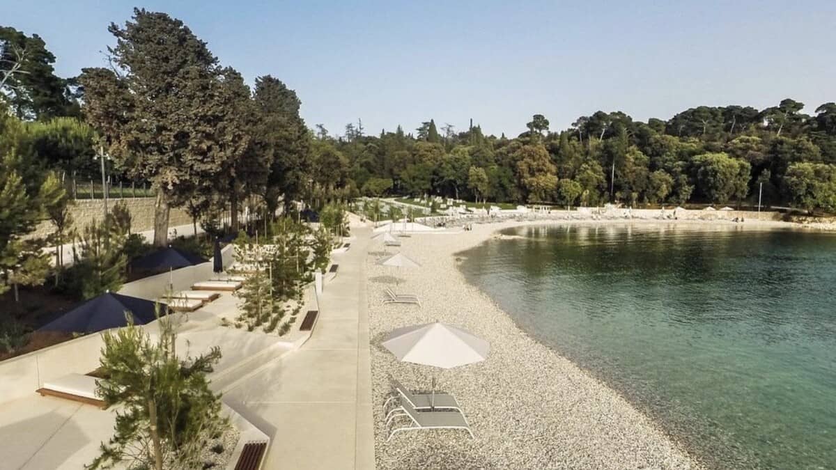 Elevated view of Mulini Beach in Rovinj, cream concrete sun-deck terraces with navy day beds and white parasols beside a pebble shore and clear green water, backed by pine forest