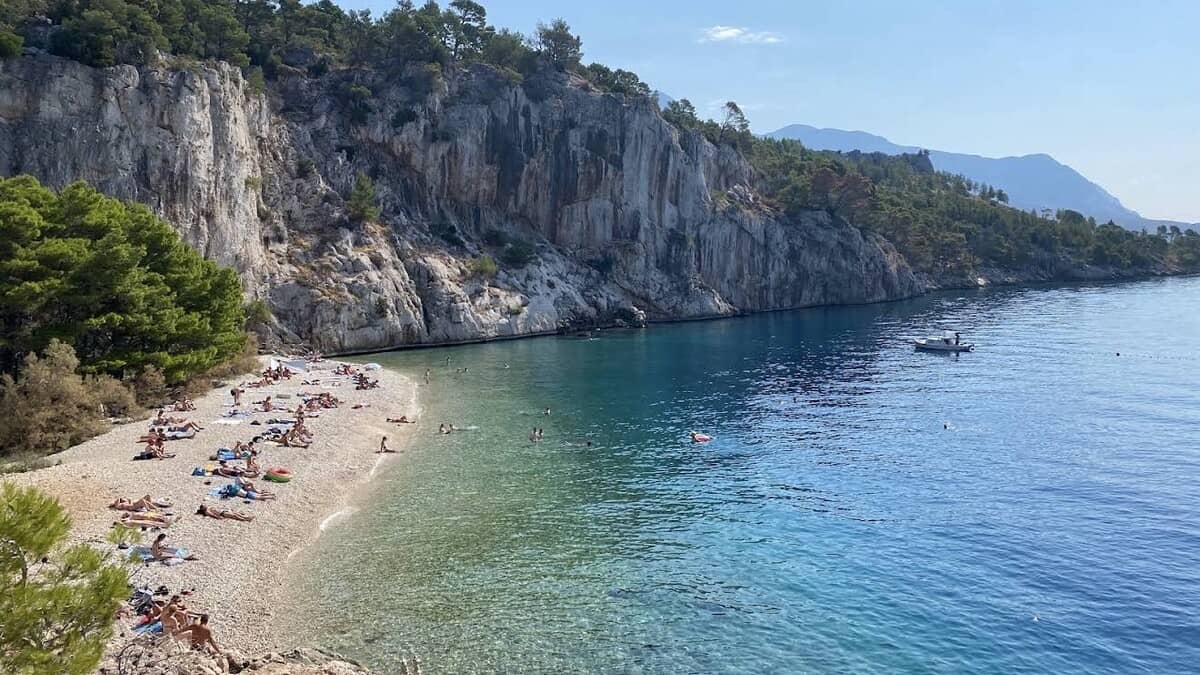 Nugal beach on the Makarska Riviera, a pebble cove of clear turquoise water with sunbathers on the shore below towering white cliffs and pine, a small boat anchored in the bay