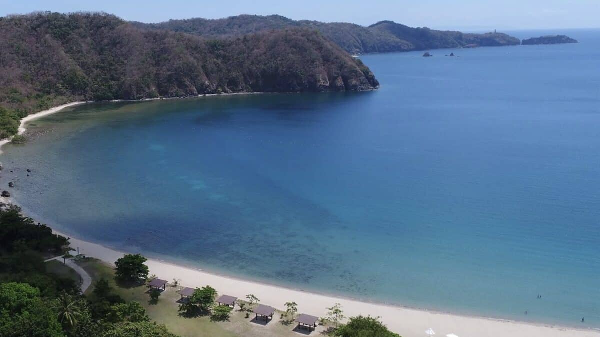 Aerial view of Pico de Loro Cove in Nasugbu, Batangas: the curved sandy shoreline of the gated resort beach with cabanas and lawns, the calm marine-reserve bay and forested headlands wrapping the cove