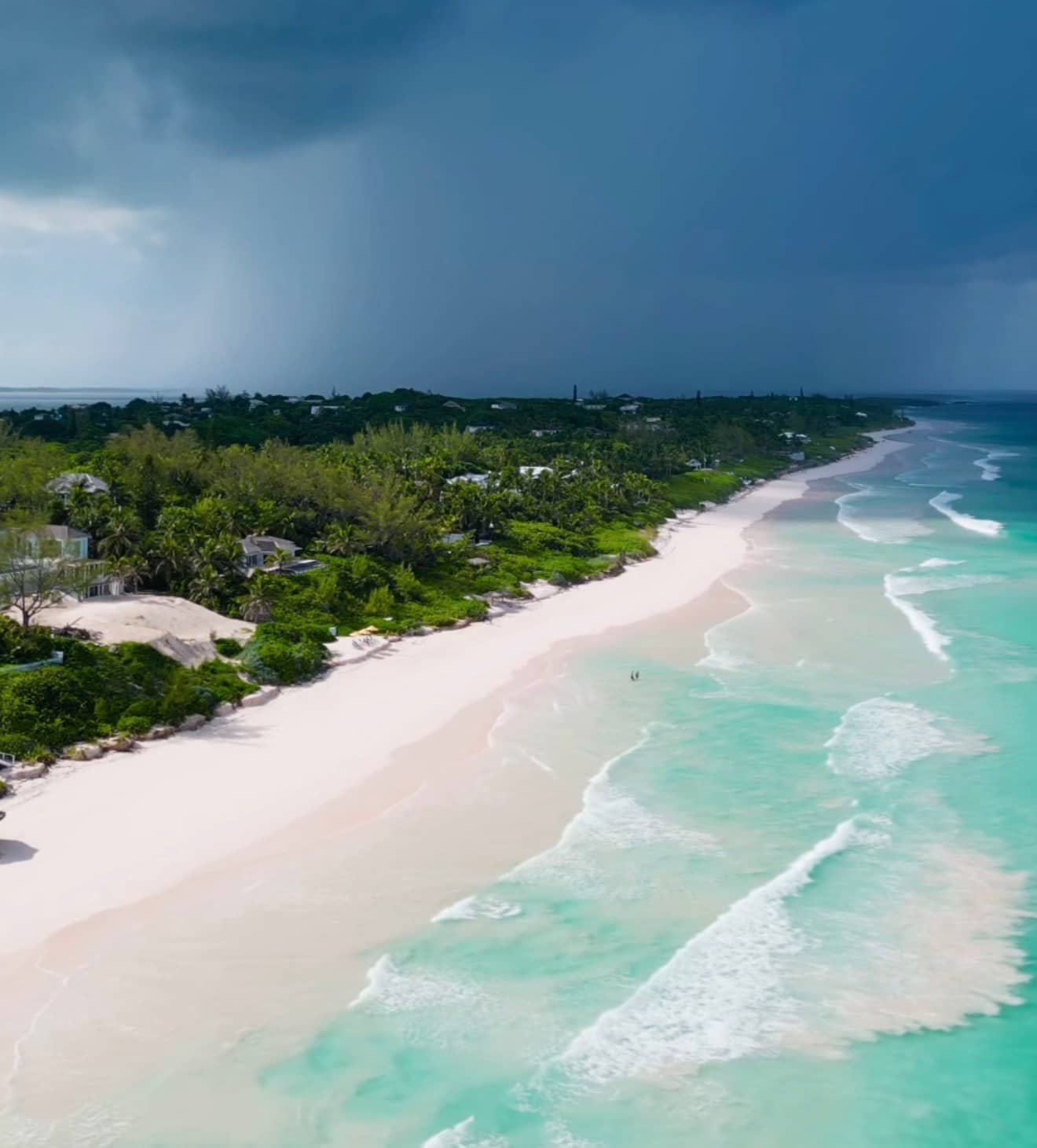 Aerial view of the Pink Sand Beach on Harbour Island in the Bahamas, showing a long curve of pale pink sand, turquoise Atlantic water, low-lying tropical vegetation and houses behind the beach, under a dramatic cloud-heavy sky