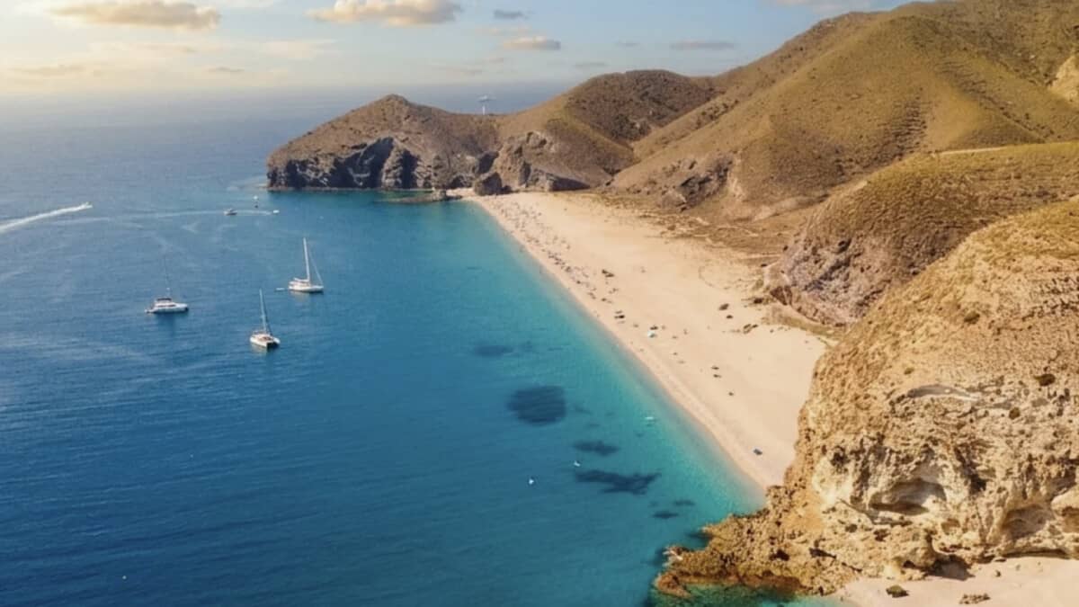 Aerial view of Playa de los Muertos near Carboneras, the long pale pebble beach curving beneath arid hills with sailing boats anchored in deep blue water