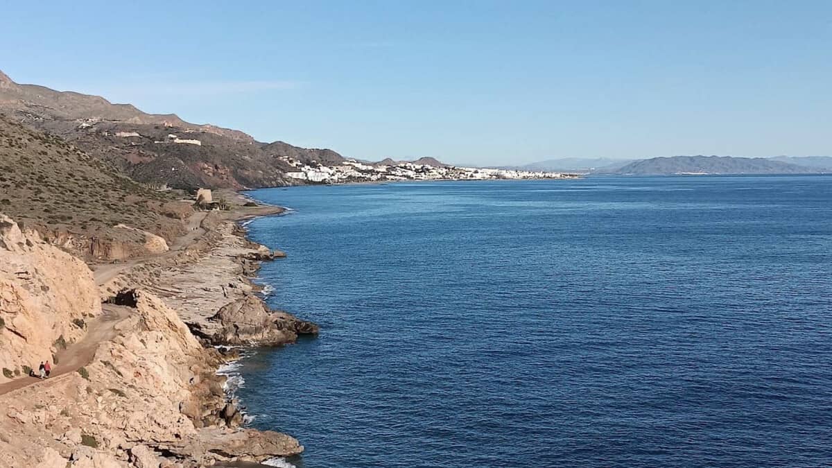 The coastal track at Macenas above the grey-sand bay, with the Torre de Macenas on the shore and the white buildings of Mojacar Playa in the distance