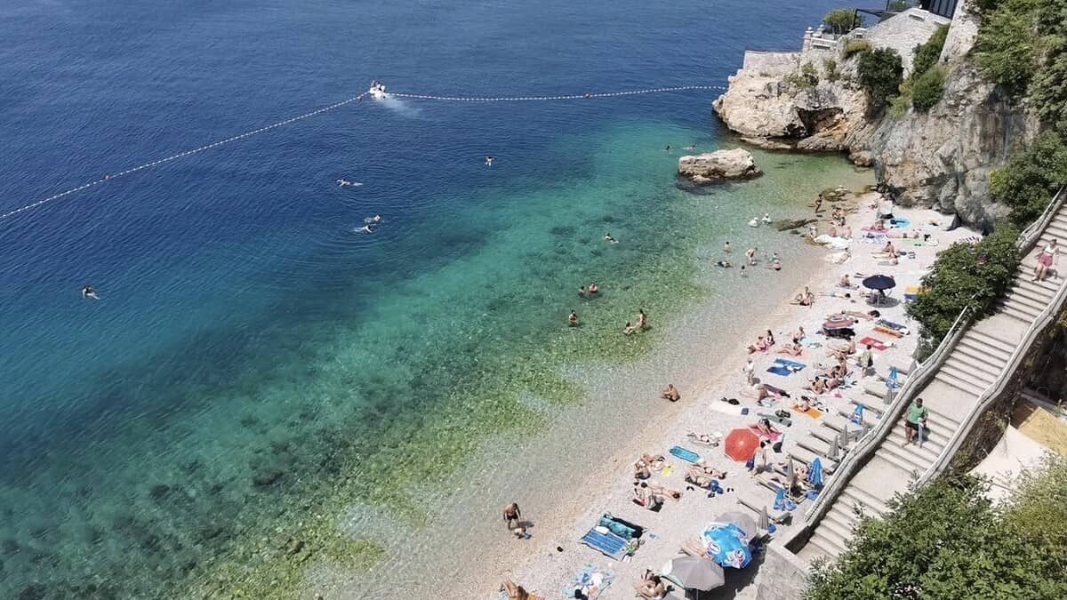 Elevated view of Sablicevo beach in Rijeka's Pecine district, a pebble cove of clear green water below the cliffs, with sunbathers, a swim-zone buoy line and the stone stairs down to the shore