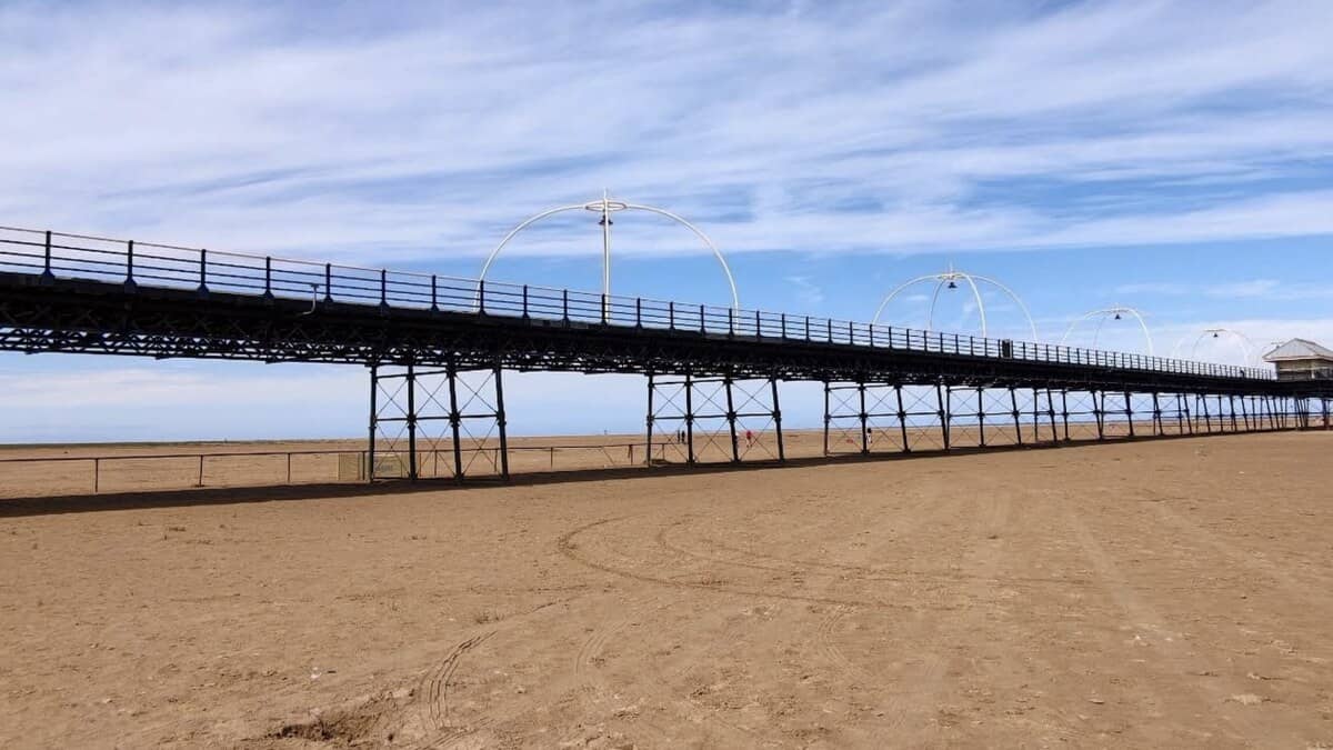 Southport's long pier striding across the vast flat sands on its iron legs and ornate arches, under a wide blue sky with the sea far out at low tide