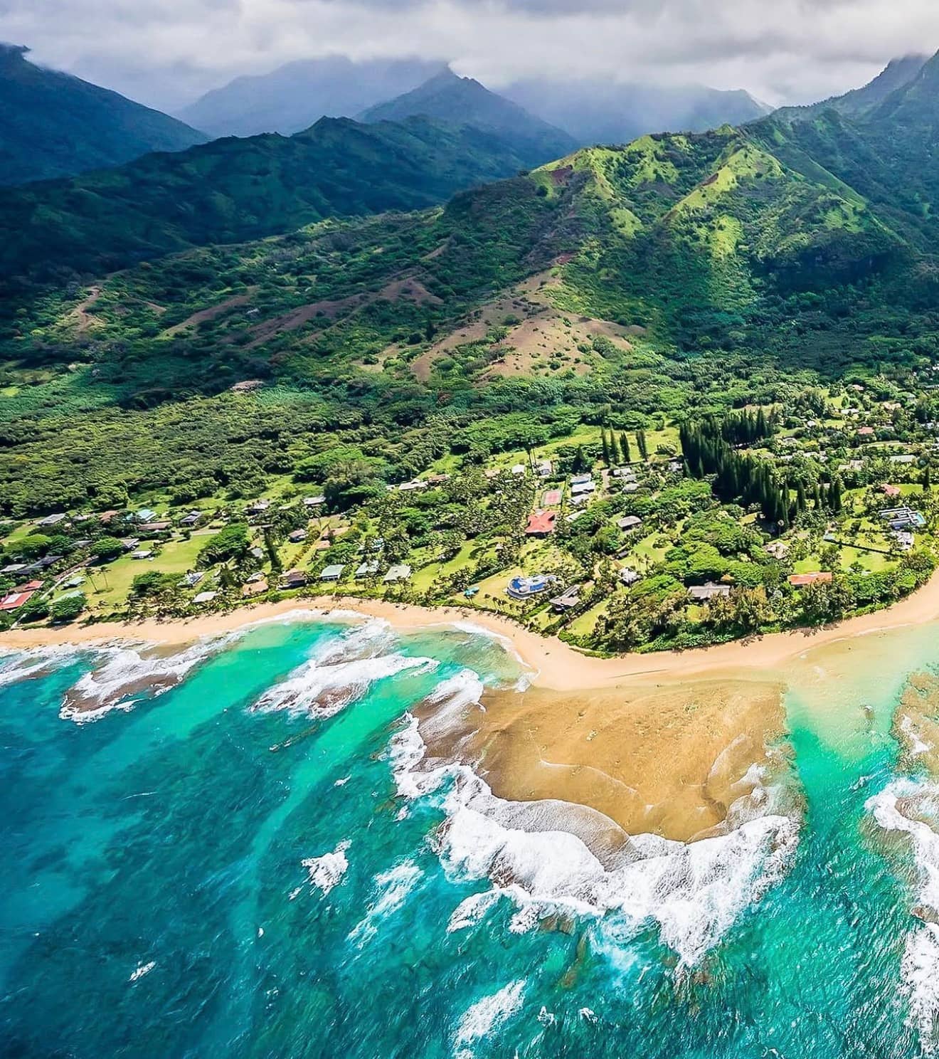 Aerial view of Tunnels Beach on Kauai's North Shore showing the curve of golden sand, the fringing reef lagoon in turquoise water, and the green Makana mountain range rising behind