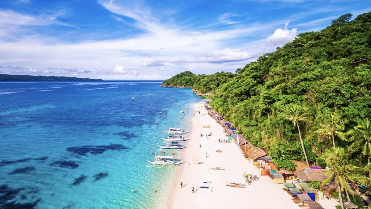 Aerial of a white-sand Boracay beach along a lush forested coast, bangka outrigger boats moored over clear turquoise water with reef patches, and people on the sand below the green hillside