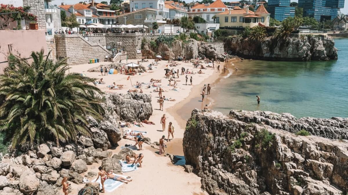 A busy golden cove beach in Cascais near Lisbon, with sunbathers, rocky outcrops and the town behind