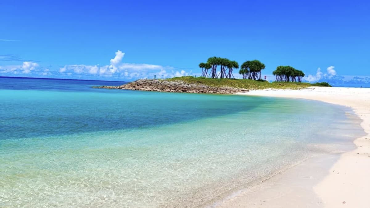 A white-sand Okinawa beach meeting graded turquoise-to-deep-blue water, with a tree-topped islet and subtropical greenery along the shore