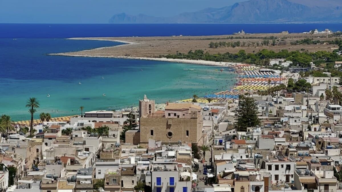 San Vito lo Capo in western Sicily seen from above, the white town spread along a wide curve of pale sand and shallow turquoise water, with a low headland and distant mountains across the bay