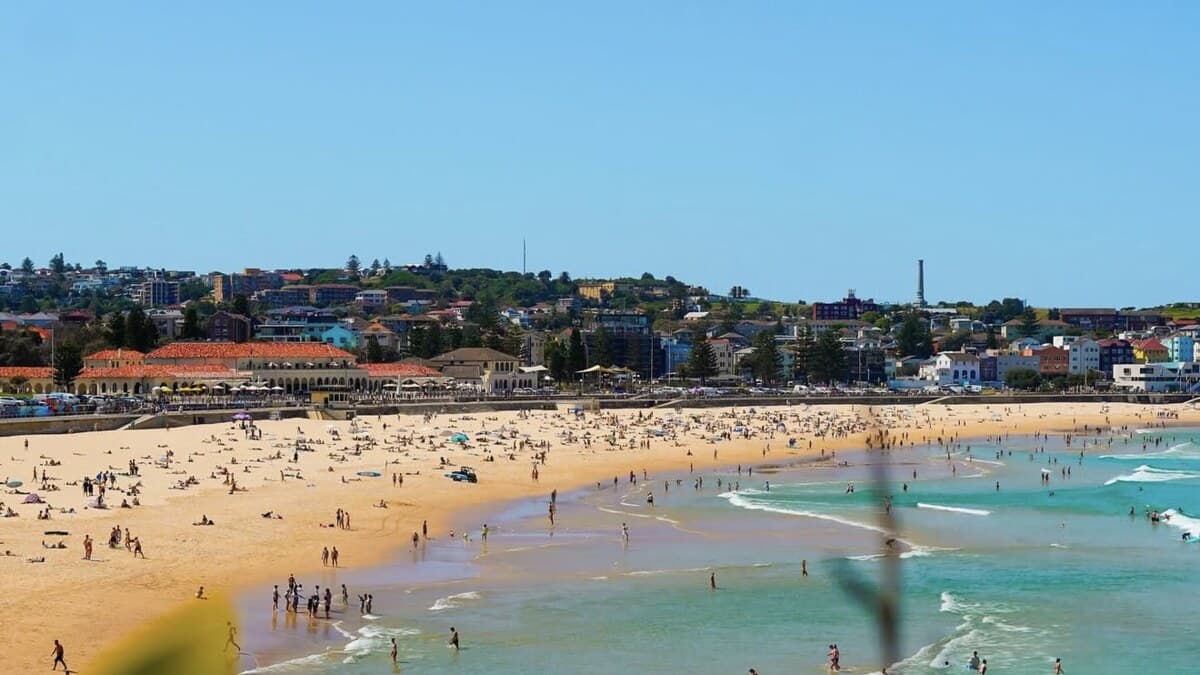Bondi Beach in Sydney seen from the southern headland, the busy golden curve of sand packed with people meeting turquoise surf, with the beachfront suburb rising behind