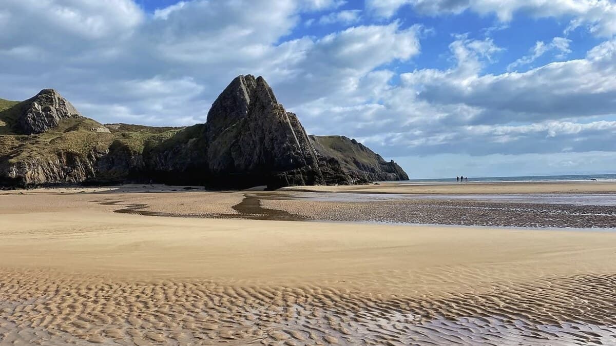 Three Cliffs Bay on the Gower, the dramatic limestone pinnacle rising above the rippled low-tide sand under a blue sky, one of the best beaches in Wales