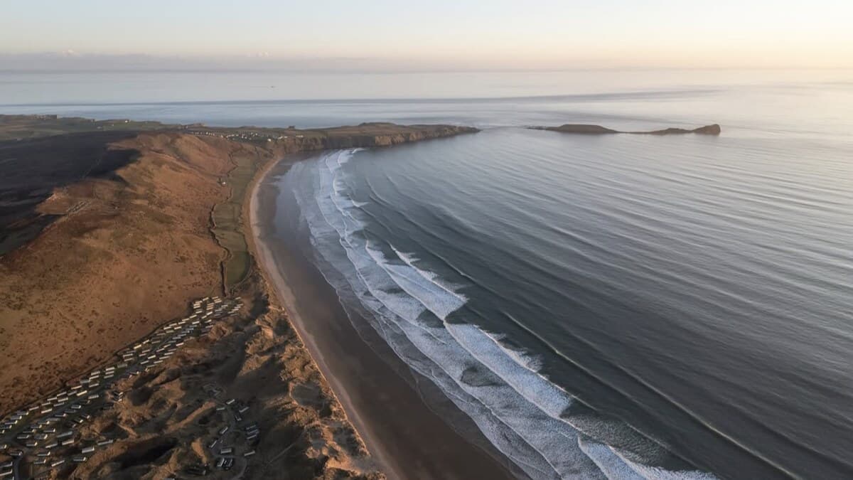 Aerial of Rhossili Bay on the Gower at dusk, the long sweep of sand and rolling surf below the down, with a clifftop caravan park and a headland beyond