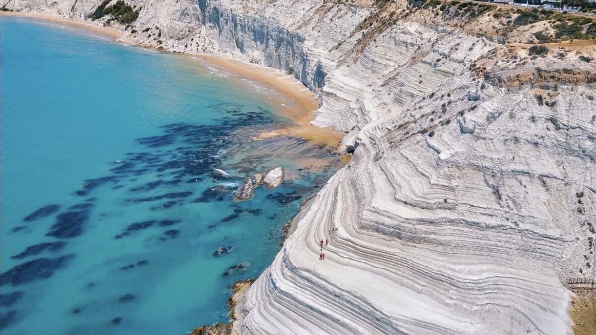 Aerial of Scala dei Turchi near Agrigento, the brilliant white marl cliffs curving in stepped layers down to turquoise sea, with a small sandy beach tucked beside them
