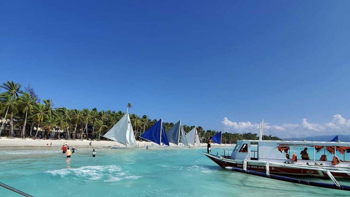 Paraw sailboats on the bright turquoise water off Boracay's busy White Beach, with swimmers near the shore