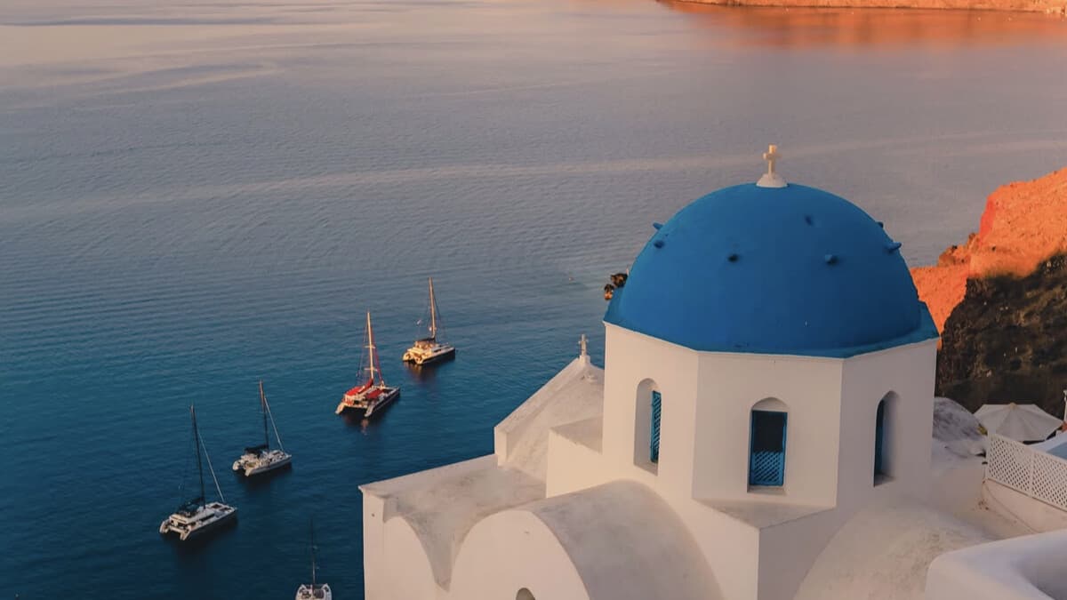 The blue-domed church at Oia on Santorini at sunset, catamarans anchored in the deep blue caldera below the whitewashed village