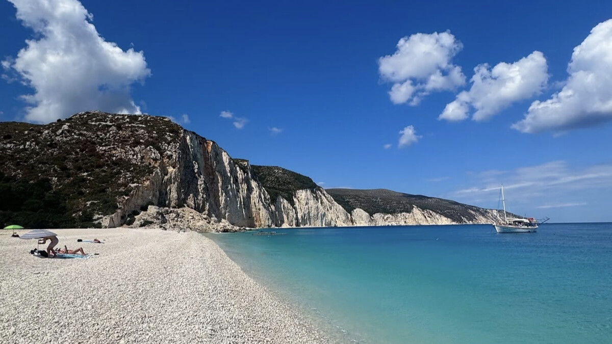 Fteri Beach on Kefalonia, a white-pebble cove beneath pale cliffs with crystal-clear turquoise water and a boat anchored offshore