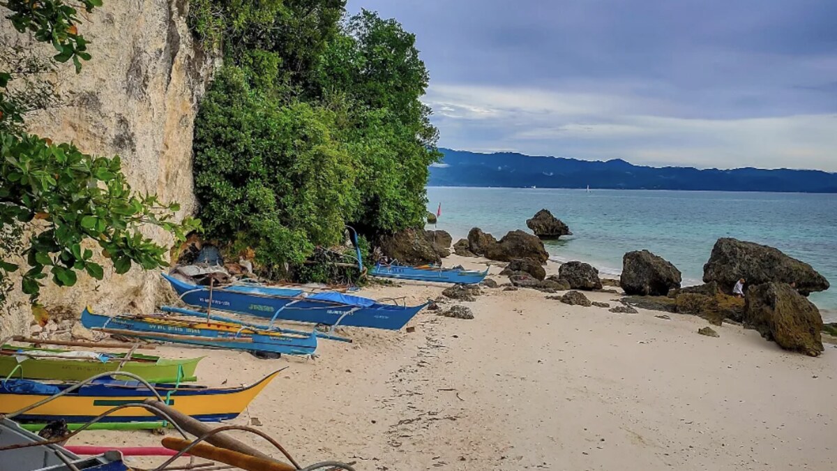 A quiet rocky corner of Diniwid Beach, Boracay, with bangka outrigger boats pulled up beneath a limestone cliff and rocks on the sand under a cloudy sky