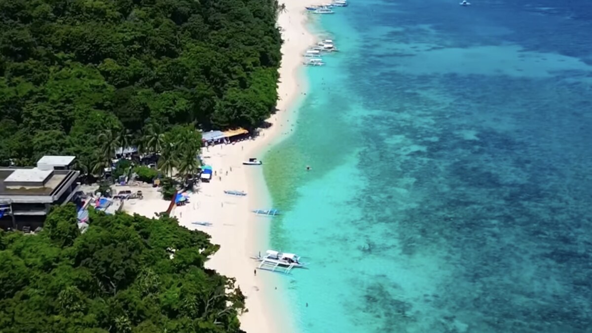 Aerial of Puka Beach on Boracay, a white-sand shore along a forested coast with bangka boats moored over clear, reef-streaked turquoise water