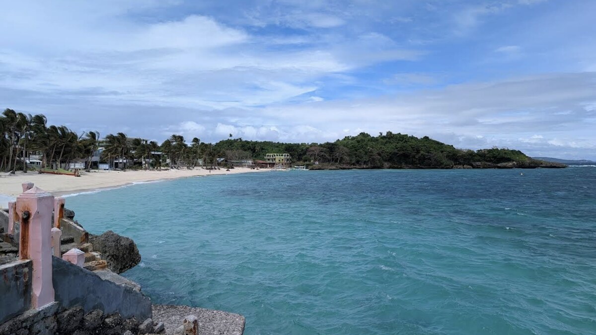 Tambisaan Beach on Boracay's quiet east coast, a calm turquoise cove with a palm-backed sandy beach and a forested headland across the water, seen from the rocky breakwater