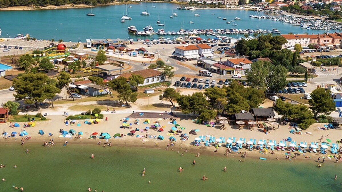 Aerial view of Bijeca beach in Medulin, a long sandy shore with swimmers wading far out in the shallow water, backed by the town and marina