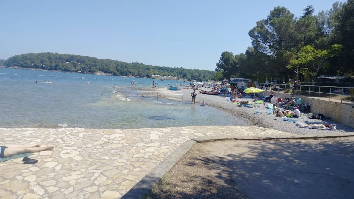 Lanterna beach near Porec, the pebble resort shore and paved promenade busy with sunbathers and umbrellas, backed by pines on the Lanterna peninsula