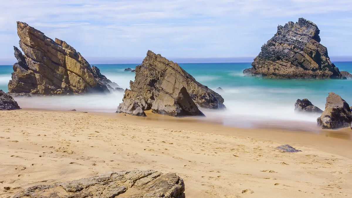 Praia da Adraga on the Sintra coast, jagged rock formations rising from the golden sand with Atlantic surf around them