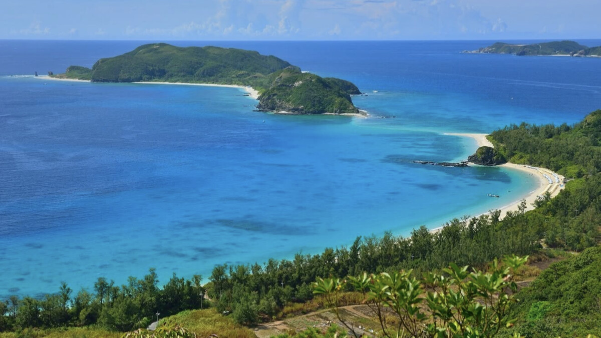 Furuzamami Beach on Zamami seen from above, the curved white-sand bay and vivid Kerama-blue water among the forested islands