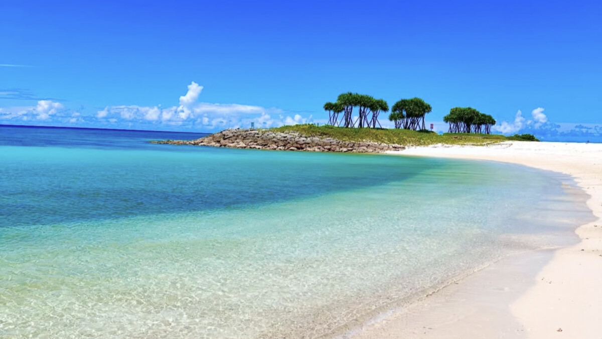A white-sand Okinawa beach meeting graded turquoise-to-deep-blue water, with a tree-topped islet and subtropical greenery along the shore
