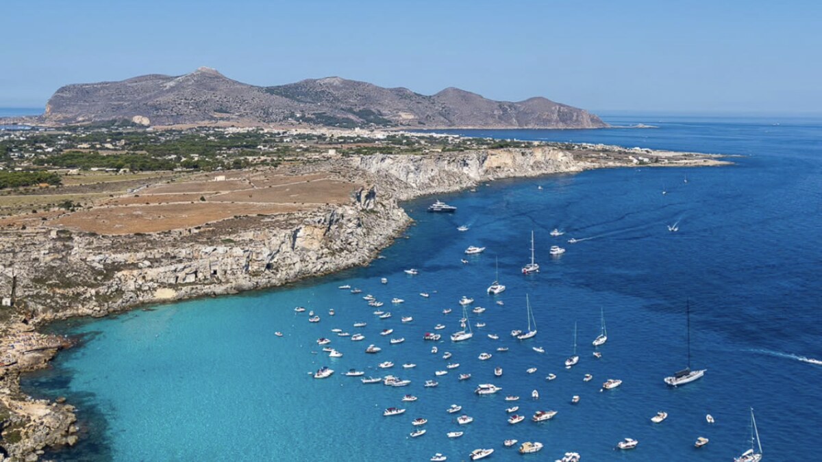 Aerial of the rocky limestone coast at Cala Rossa on Favignana, boats moored over vivid turquoise water with the island and distant hills behind