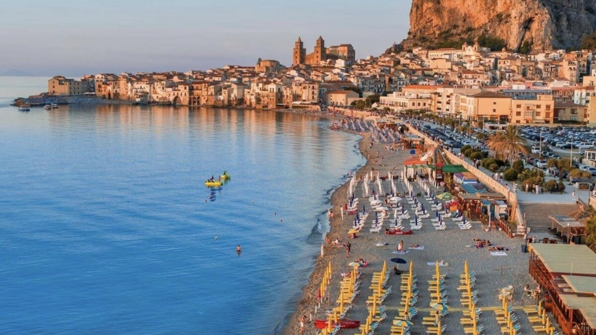 Cefalu in northern Sicily at golden hour, rows of sun loungers on the town beach below the medieval old town and Norman cathedral, with the La Rocca crag behind