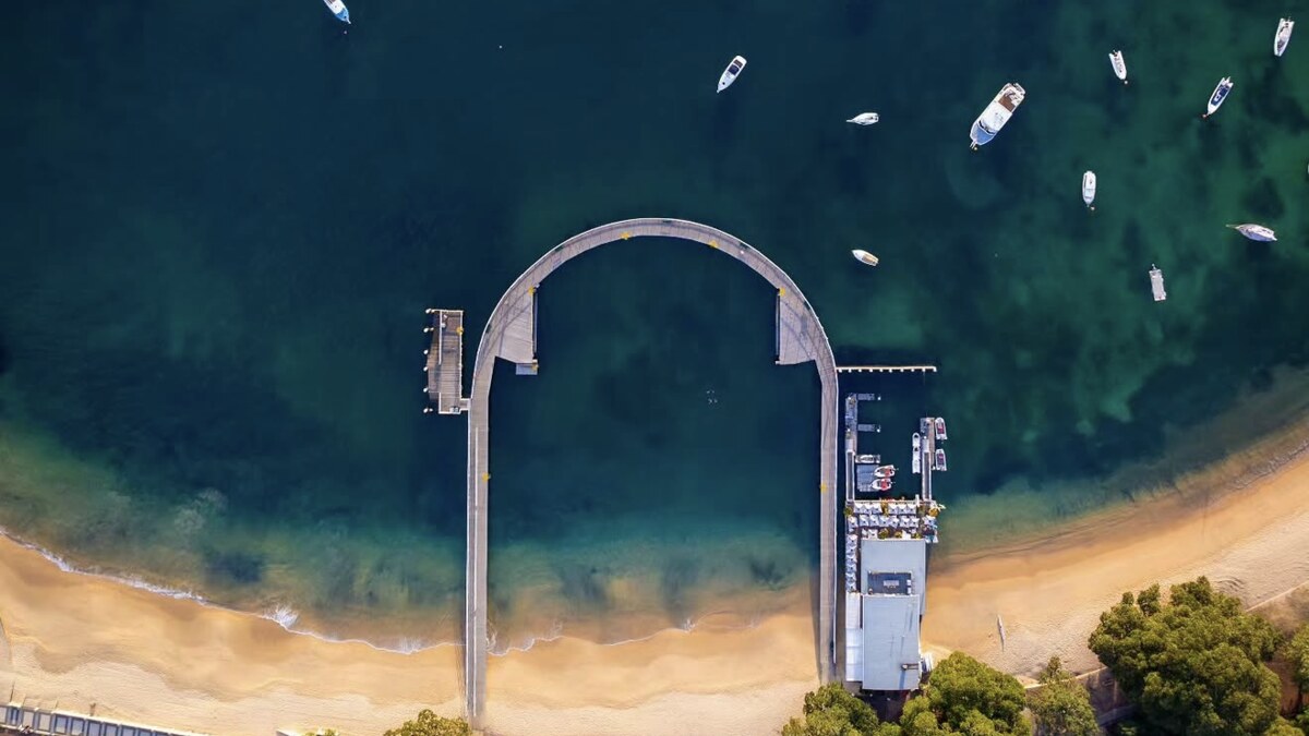 Aerial looking straight down on Balmoral Beach in Sydney Harbour, the arched footbridge to Rocky Point Island, moored boats on the calm clear water and the sandy beach below