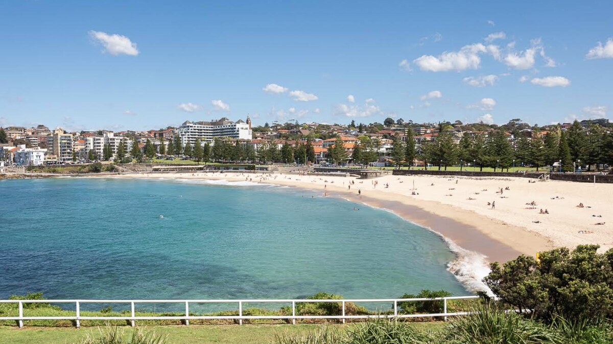 Coogee Beach in Sydney, a sheltered curve of golden sand and calm turquoise water seen from the grassy headland reserve, with the suburb rising behind