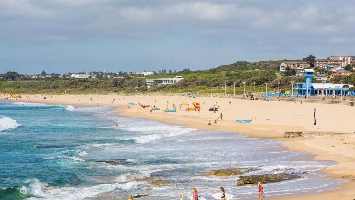 Maroubra Beach in Sydney, a long broad surf beach with waves rolling in and surfers on the sand, backed by a low green headland and the surf club