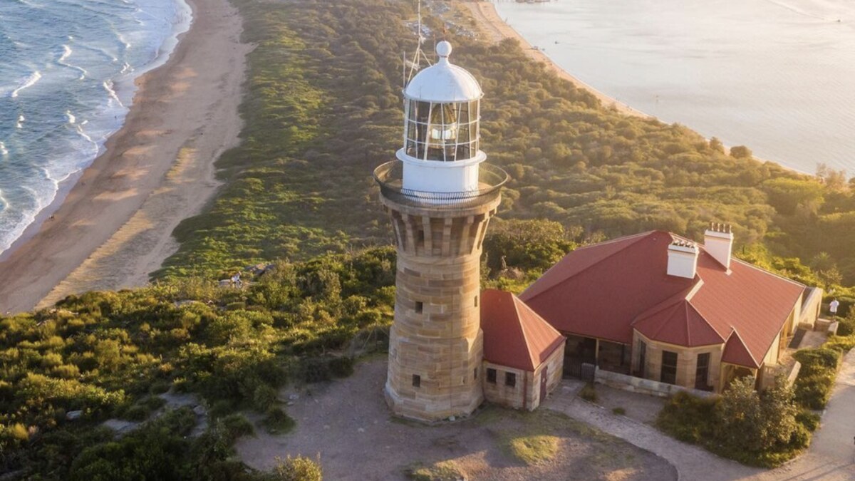 Aerial of the Barrenjoey lighthouse and keeper's cottage above Palm Beach in Sydney's far north at golden hour, the ocean surf beach on one side and the calm Pittwater on the other