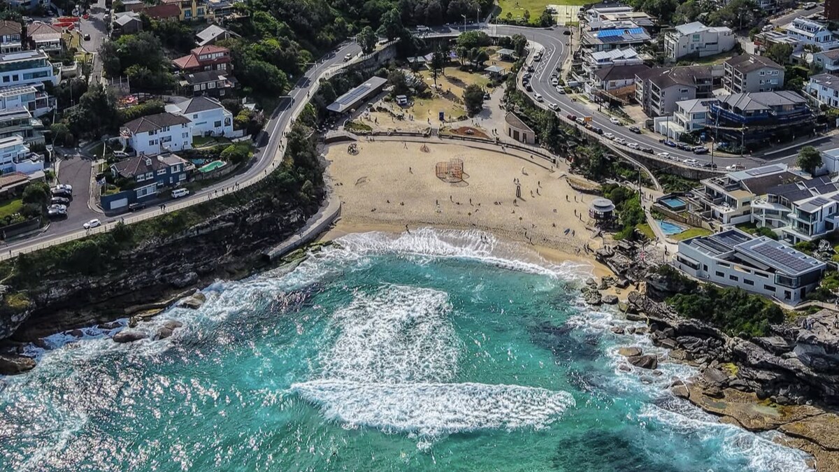 Aerial of Tamarama Beach in Sydney, a small sandy cove wedged between rocky headlands with surf rolling in, houses and a grassy park wrapped around it